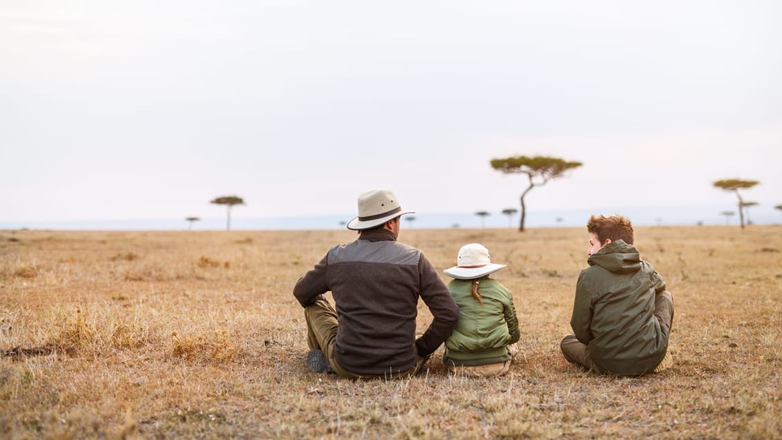 Three travellers, one a child, sitting on an open plain with acacias behind