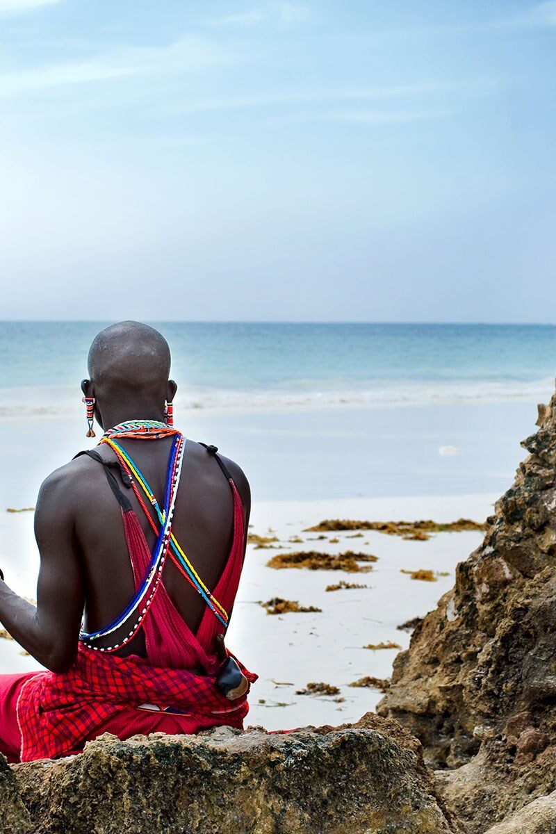 A Maasai man with spear and shield sitting on rocks above a turquoise sea, Kenya