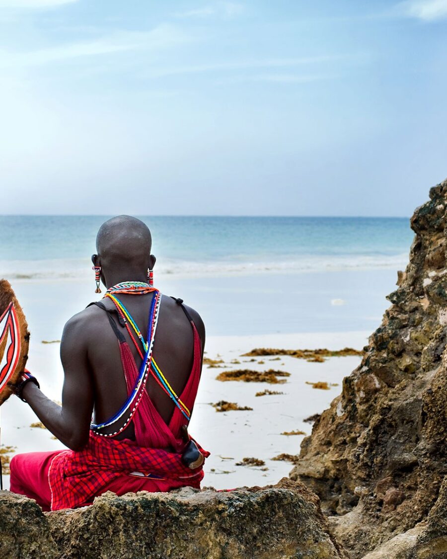 A Maasai man with spear and shield sitting on rocks above a turquoise sea, Kenya